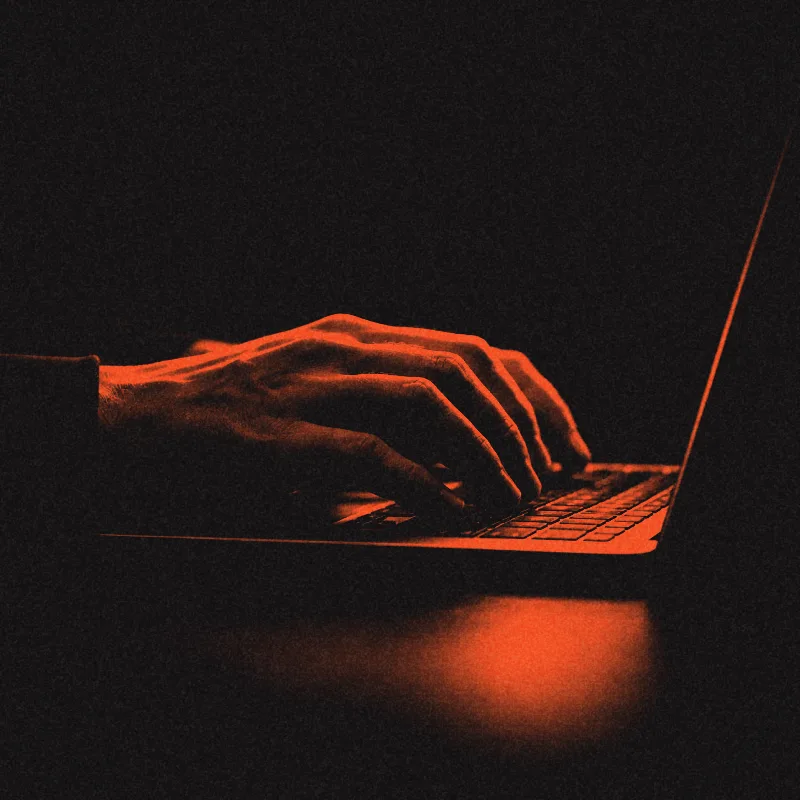 Hands typing on a laptop, lit in ember against black